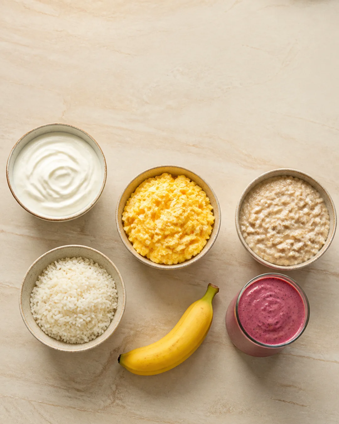 Soft foods to eat after a root canal arranged on a light counter: yogurt, scrambled eggs, oatmeal, a bowl of rice, a banana, and a smoothie